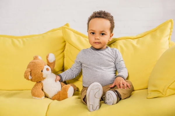 Adorable enfant afro-américain tenant ours en peluche et regardant la caméra tout en étant assis sur le canapé jaune — Photo de stock