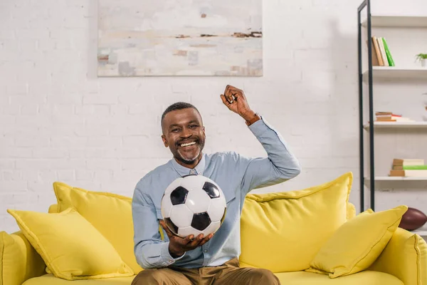 Homem americano africano alegre segurando bola de futebol e olhando para a câmera enquanto sentado no sofá em casa — Fotografia de Stock