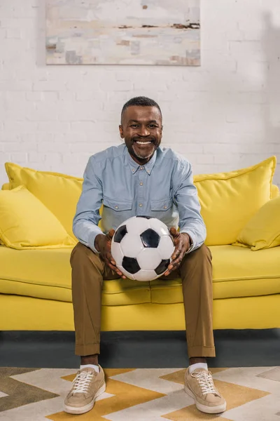 Sorrindo homem americano africano sentado no sofá e segurando bola de futebol — Fotografia de Stock