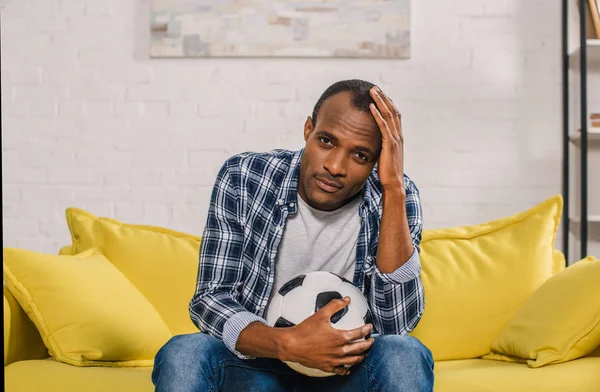 Sconvolto uomo afroamericano in possesso di palla da calcio e guardando la fotocamera mentre seduto sul divano a casa — Foto stock