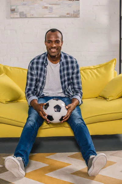 Fröhlicher afrikanisch-amerikanischer Mann hält Fußballball in der Hand und lächelt in die Kamera, während er auf der Couch sitzt — Stockfoto