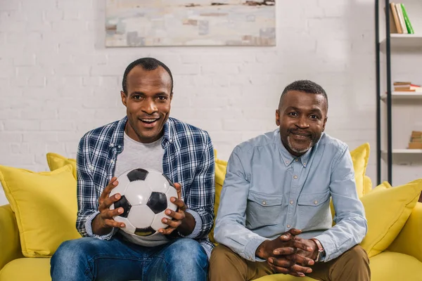 Feliz afroamericano padre con adulto hijo celebración fútbol pelota y sonriendo a la cámara en casa - foto de stock