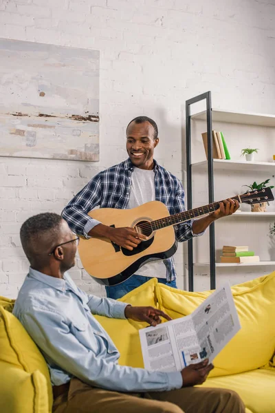 Feliz hombre mayor con periódico e hijo adulto con la guitarra sonriendo el uno al otro en casa - foto de stock