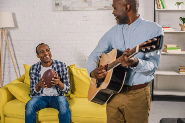 Cropped shot of senior man playing acoustic guitar and looking at smiling adult son holding rugby ball at home — Stock Photo
