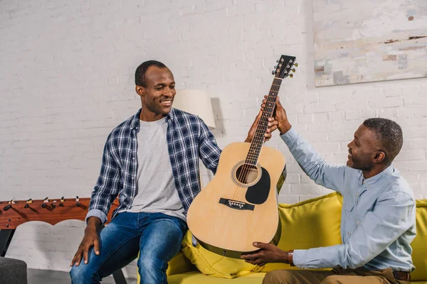 Happy senior man giving guitar to smiling adult son at home — Stock Photo