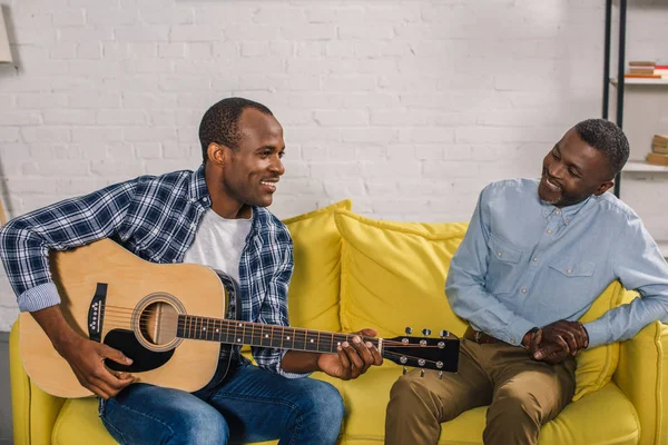 Sorrindo homem sênior olhando para o filho adulto tocando guitarra em casa — Fotografia de Stock