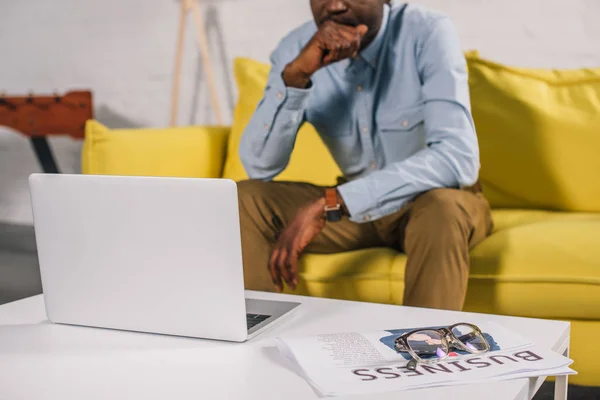 Vue rapprochée de l'ordinateur portable, des lunettes et du journal sur la table et de l'homme afro-américain assis sur le canapé derrière — Photo de stock