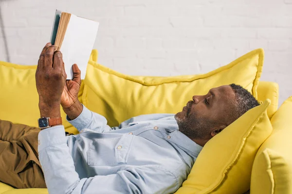 Vista lateral del hombre afroamericano mayor acostado en un sofá y leyendo un libro - foto de stock
