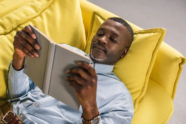 Senior african american man lying on sofa and reading book — Stock Photo