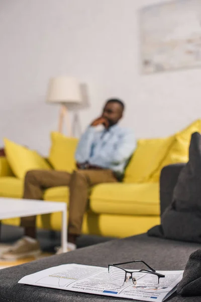 Nahaufnahme von Brille und Zeitung auf der Couch und ein älterer afrikanisch-amerikanischer Mann sitzt dahinter — Stockfoto