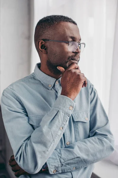 Pensive senior african american man in eyeglasses standing with hand on chin and looking at window — Stock Photo