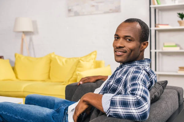 Guapo joven afroamericano hombre sentado en el sofá y sonriendo a la cámara - foto de stock