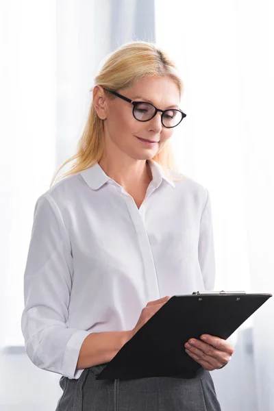 Mature female therapist in eyeglasses writing in clipboard — Stock Photo