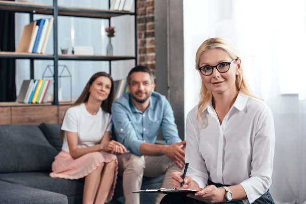 Consejera femenina escribiendo en portapapeles y mirando a la cámara mientras la familia feliz sentado detrás en el sofá en la oficina - foto de stock