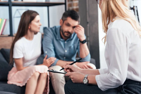 Selektiver Fokus der weiblichen Beraterin, die in die Zwischenablage schreibt, während Frau gestressten Mann auf Therapiesitzung umarmt — Stockfoto