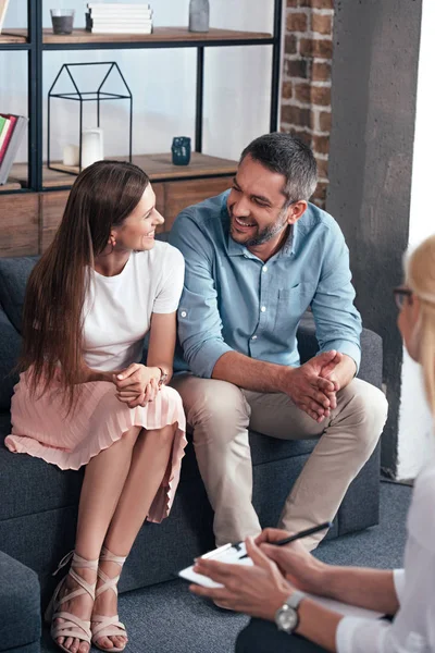 Lächelndes Paar, das sich anschaut und bei einer Therapiesitzung von einer Beraterin im Büro sitzt — Stockfoto