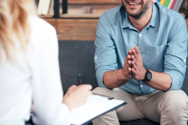 Image recadrée d'une conseillère écrivant dans un presse-papiers et d'un homme souriant tenant les paumes de la main ensemble pendant une séance de thérapie au bureau — Photo de stock