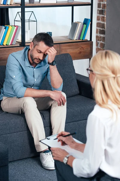 Depressed man sitting on sofa while female psychiatrist writing in clipboard at office — Stock Photo