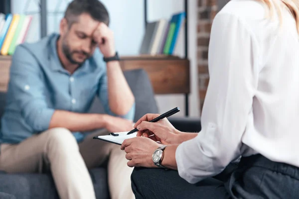 Cropped image of female counselor writing in clipboard while depressed male patient sitting on sofa at office — Stock Photo