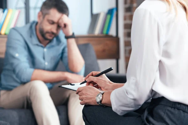 Imagen recortada de la escritura del consejero femenino en portapapeles mientras paciente masculino deprimido sentado en el sofá en la oficina - foto de stock