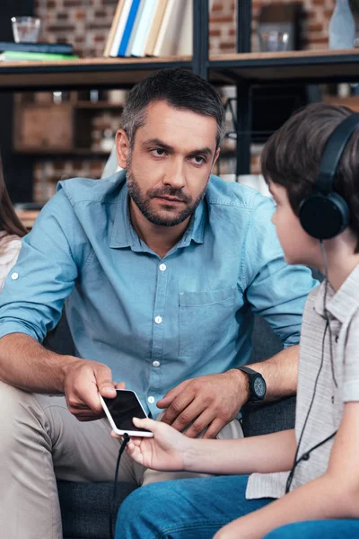 Серьезный отец забирает смартфон у маленького сына в наушниках — стоковое фото
