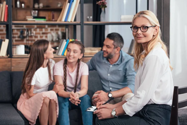 Souriant femme psychiatre regardant caméra tandis que la famille heureuse avec fille en thérapie session — Photo de stock