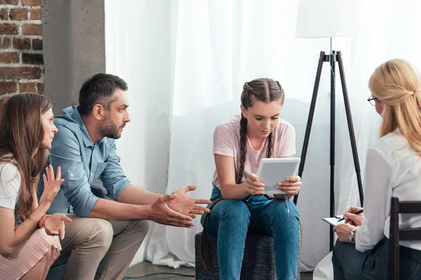 Parents gesturing by hands and trying to talk with teenage girl using digital tablet on therapy session by female counselor in office — Stock Photo