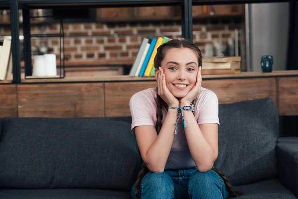 Sorridente ragazza adolescente con le mani sulle guance guardando la fotocamera e seduto sul divano a casa — Foto stock