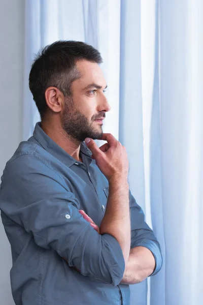 Side view of thoughtful man with hand on chin in front of curtains at home — Stock Photo