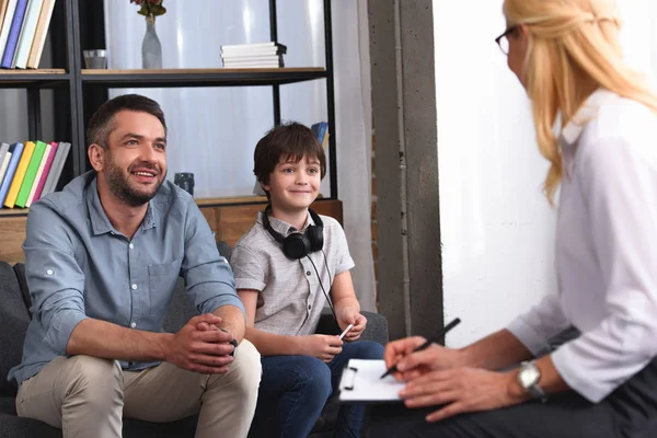 Side view of female counselor writing in clipboard while father and son with headphones sitting on therapy session in office — Stock Photo