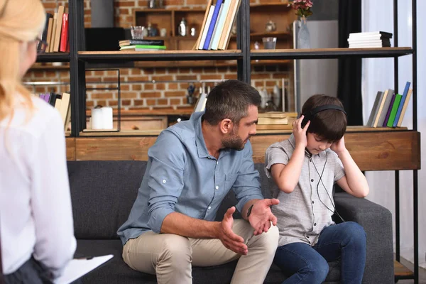 Батько з широкими руками розмовляє з маленьким сином в навушниках на сесії сімейної терапії жінкою-консультантом — стокове фото