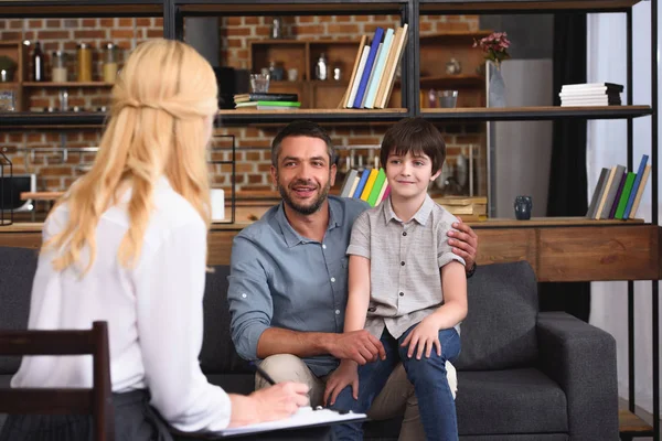 Vista posteriore di consigliere donna che scrive negli appunti mentre padre e figlio sono seduti in sessione di terapia — Foto stock