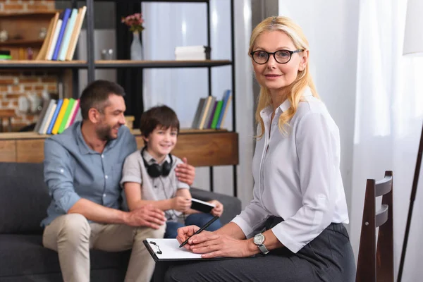 Beraterin schreibt in Klemmbrett und blickt in Kamera, während Vater und Sohn auf Therapiesitzung sitzen — Stockfoto