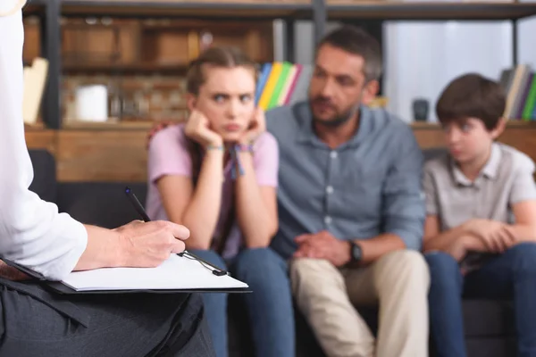 Immagine ritagliata di consigliere donna che scrive negli appunti mentre la famiglia siede sul divano in sessione di terapia — Foto stock