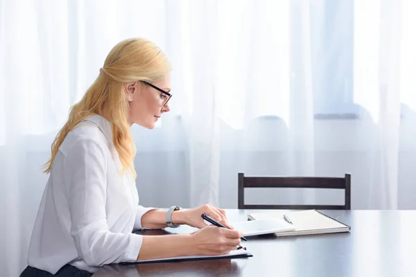 Seitenansicht einer Psychiaterin mit Brille, die auf den digitalen Tablet-Bildschirm am Tisch im Büro blickt — Stockfoto