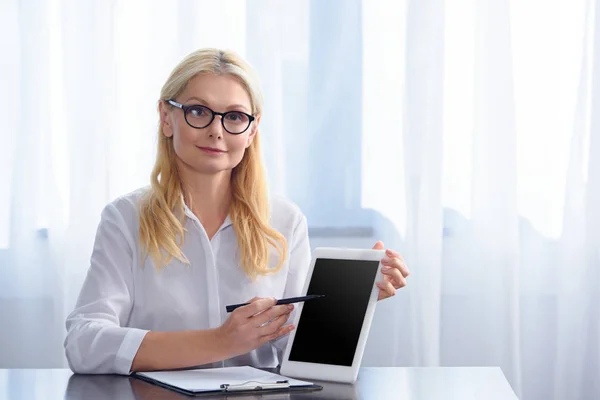 Улыбающаяся женщина-консультант в очках, указывающая на цифровой планшет с пустым экраном за столом в офисе — стоковое фото