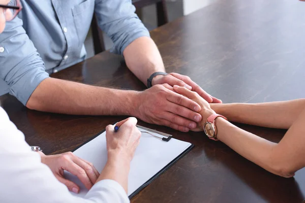 Cropped image of couple holding hands of each other while female counselor writing in clipboard at table in office — Stock Photo