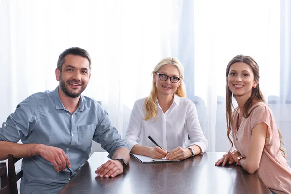 Coppia sorridente e consulente femminile che scrivono negli appunti a tavola in ufficio — Foto stock