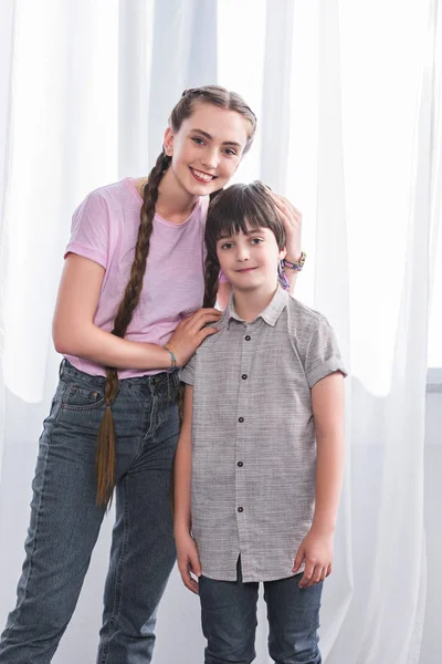 Feliz adolescente chica abrazando pequeño hermano y mirando a la cámara en casa - foto de stock