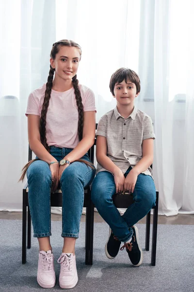 Улыбающаяся девочка-подросток с братом, сидящим на стульях и смотрящим в камеру дома — стоковое фото