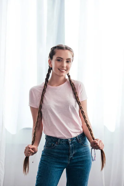 Feliz adolescente mirando a la cámara y sosteniendo sus propias trenzas delante de las cortinas en casa - foto de stock