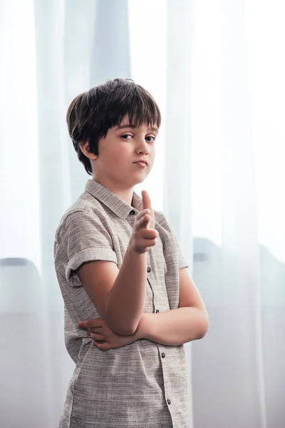 Niño pequeño haciendo el arma de mano y mirando a la cámara delante de las cortinas en casa - foto de stock