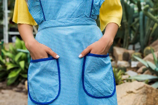 Cropped shot of woman in apron standing with hands in pockets in greenhouse — Stock Photo