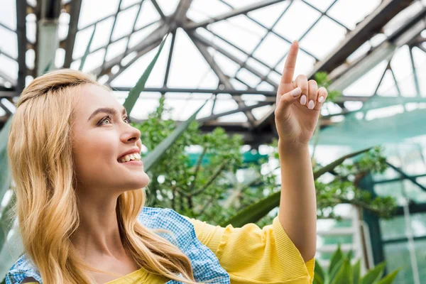 Schöne lächelnde blonde Frau zeigt mit dem Finger nach oben im Gewächshaus — Stockfoto