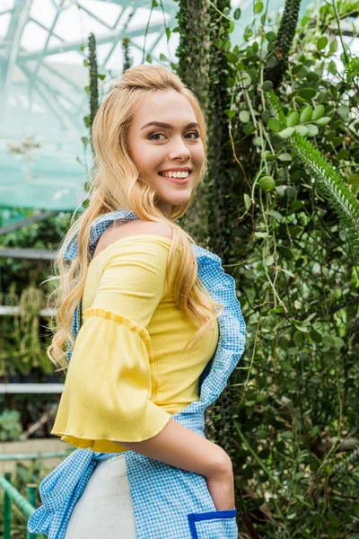 Vista lateral de la hermosa chica en delantal sonriendo a la cámara mientras está de pie cerca de plantas verdes - foto de stock