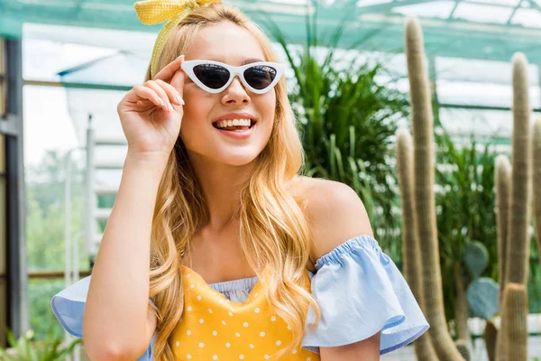 Belle fille blonde heureuse dans les lunettes de soleil et tablier souriant dans la serre — Photo de stock