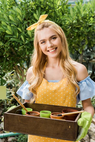 Beautiful young female gardener holding box with flower pots and smiling at camera — Stock Photo