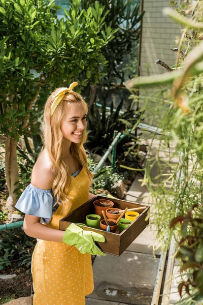 Vista de alto ângulo de bela mulher sorridente segurando caixa com vasos de flores e olhando para longe em estufa — Fotografia de Stock