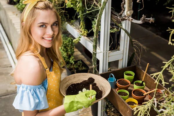 Vista de ángulo alto de la hermosa mujer joven sosteniendo tazón con tierra y sonriendo a la cámara en invernadero - foto de stock