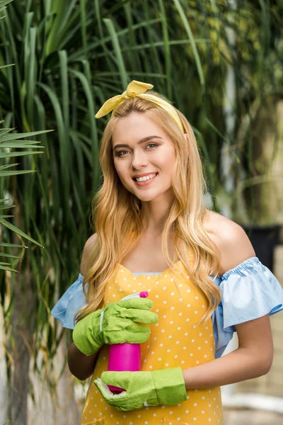 Bella giovane donna in guanti di gomma in possesso di spruzzatore e sorridente alla fotocamera in serra — Foto stock